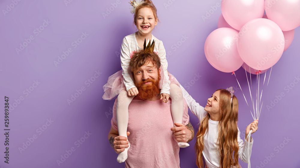 Fototapeta premium Busy redhead bearded man gives piggyback to little daughter, older sister stands near, holds air balloons, stand over purple background, celebrate holidays together. Family and celebration concept