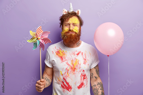 Photos Studio shot of dissatisfied redhead man holds toy windmill and helium balloon, has face dirty with yellow watercolours, ginger hair and beard, isolated over purple background