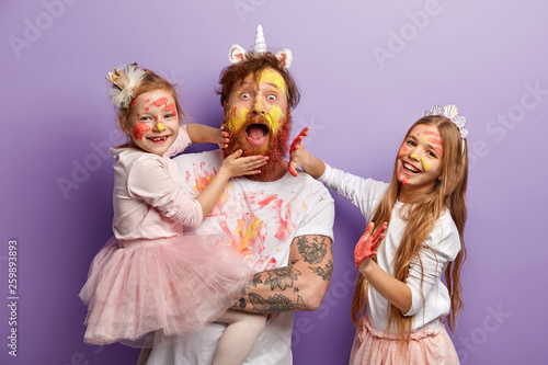 Schilderij op canvas Emotional bearded busy father spends time with two naughty daughters who leave palm prints on his beard and clothes, learn how to paint, dressed in festive clothes, stand indoor