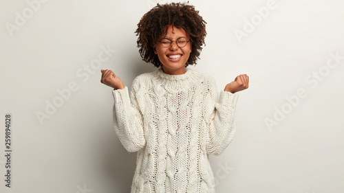 Half length shot of optimistic female feels like winner, raises hands in fists, dressed in white knitted sweater, closes eyes from pleasure isolated over white background. People, success, joy concept