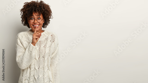 Cheerful dark skinned female shows hush sign, keeps fore finger over lips, smiles happily, tells pleasant secret to close friend, wears white winter outfit, stands in studio, blank space for your text