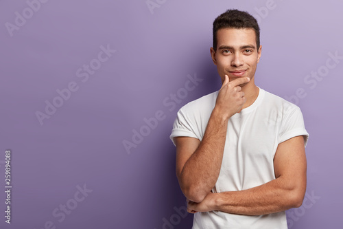 Half length shot of handsome European man holds chin, looks with satisfied confident expression directly at camera, wears white t shirt, poses in studio over purple wall, free space left aside