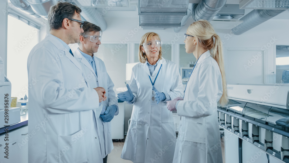 Team of Research Scientists Have Meeting, They Have Discussion While Standing in the Middle of Laboratory. People in Innovative Modern Lab Doing Genetics and Pharmaceutical Studies. Team of Research Scientists Have Meeting, They Have Discussion While Standing in the Middle of Laboratory. People in Innovative Modern Lab Doing Genetics and Pharmaceutical Studies.