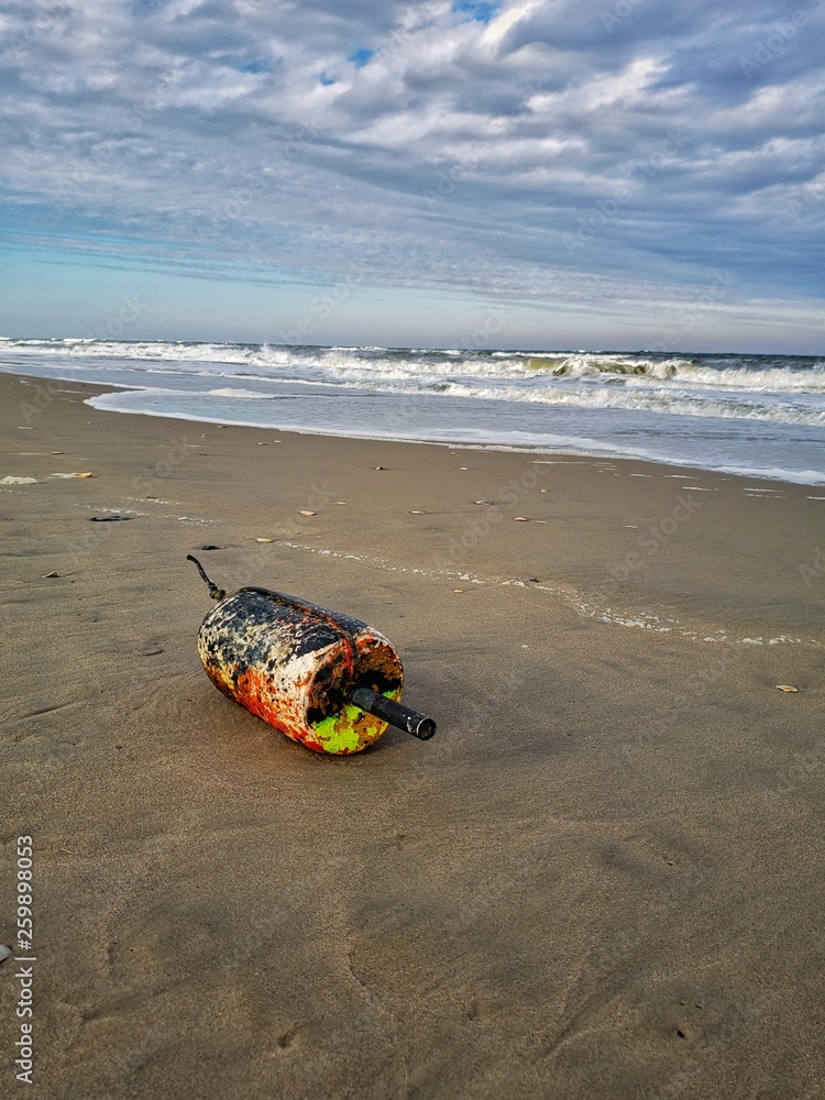 Obraz premium Float from a crab trap washed ashore on a sandy beach. Waves crash onto the shore in the background and cloudy sky. Fishing industry trash and debris. Coastal pollution.