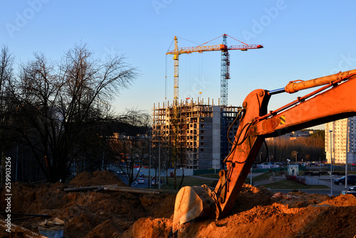 Excavator bucket in the ground at a construction site against the backdrop of a sunset and cranes