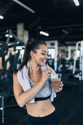 Young attractive woman after successful workout in modern fitness gym holding glass of protein shake and drinks with drinking straw.