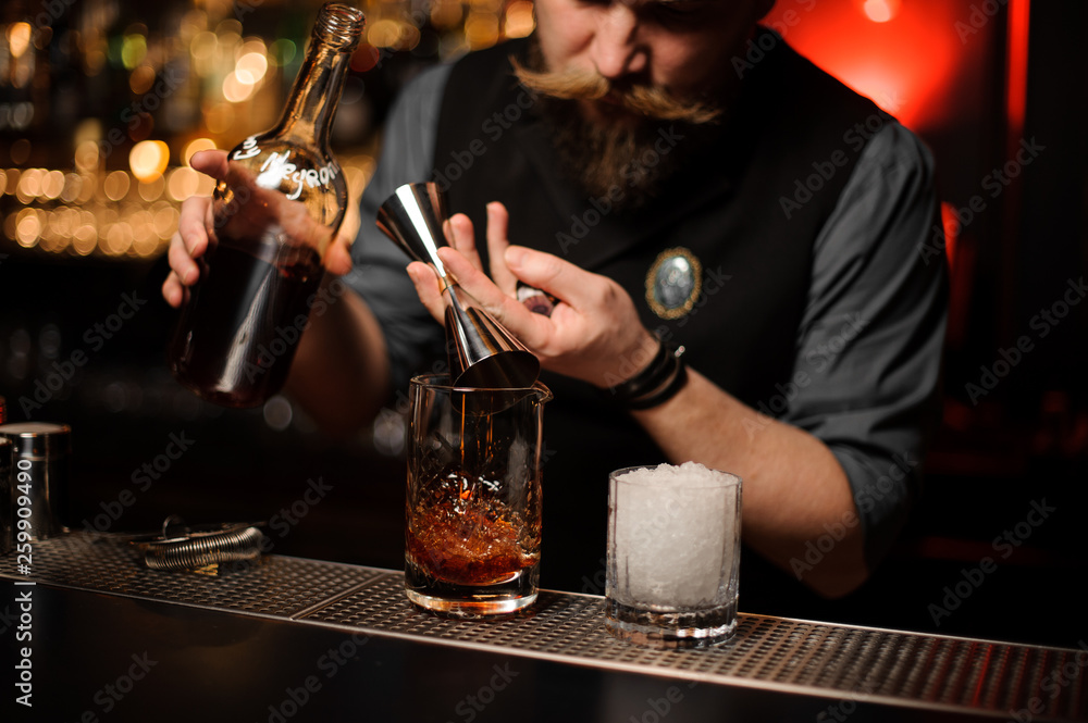 Male bartender pouring a delicious brown cocktail from the measuring glass cup through the strainer
