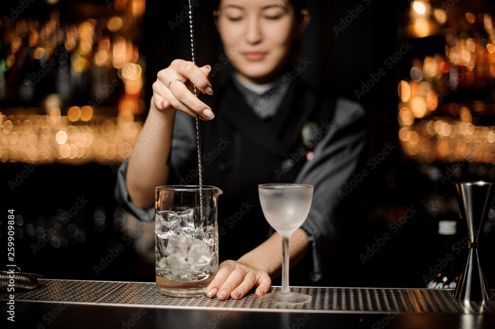 Bartender girl stirring an ice cubes with a steel spoon in the measuring glass cup