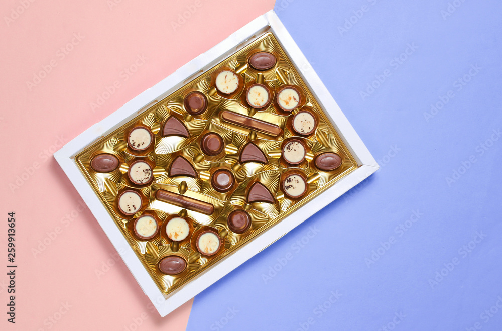 Open box with golden tray of variety of chocolate candies on plue pink pastel background. Top view, minimalism. Open box with golden tray of variety of chocolate candies on plue pink pastel background. Top view, minimalism.