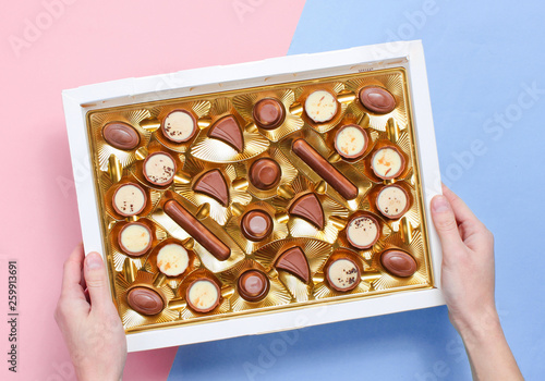 Girl holding her hands a box of chocolates in a golden tray on a pastel background. Top view.