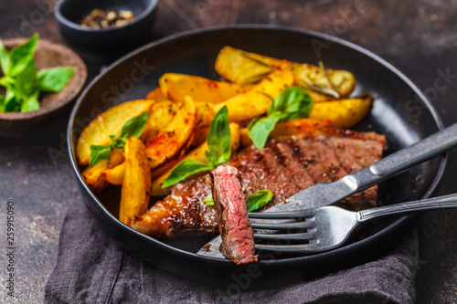 Grilled beef steak with potatoes and basil in a black plate on dark background.