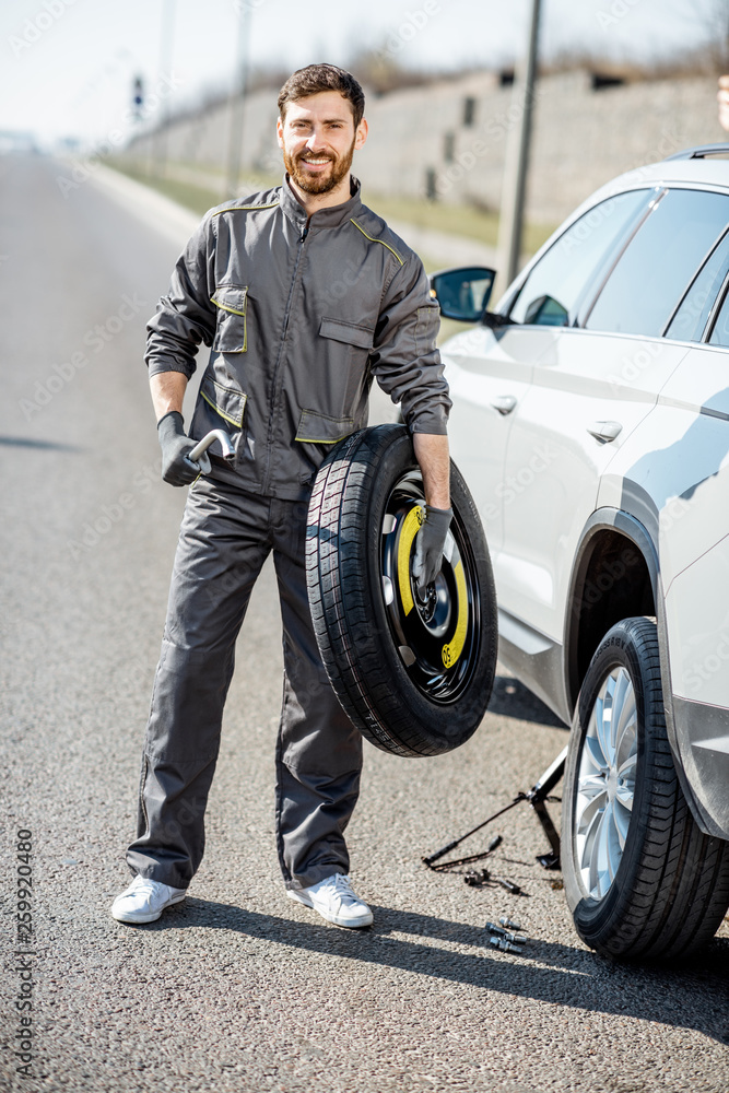 Portrait of a handsome road assistance worker in uniform standing with spare wheel near the broken car on the highway Portrait of a handsome road assistance worker in uniform standing with spare wheel near the broken car on the highway