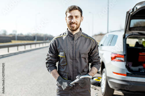 Portrait of a handsome road assistance worker in uniform standing near the broken car on the highway