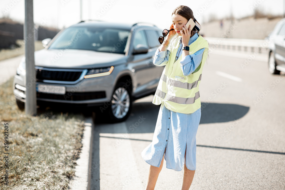 Embarrassed woman calling road assistance standing near the car during the road accident on the highway