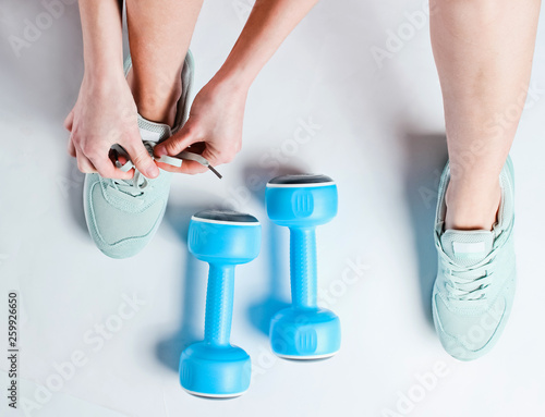 Legs of young sports woman, plastic dumbbells. Woman tying shoelace of sports shoes on a white background. Top view. Minimalism