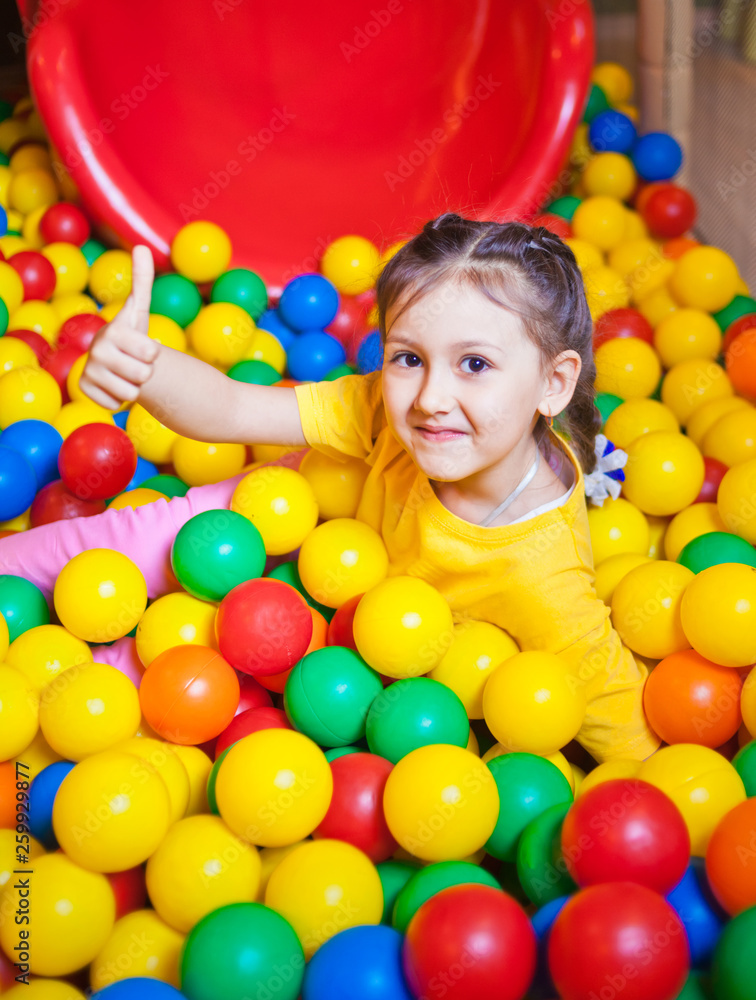 Happy little girl playing and having fun at kindergarten with colorful balls and show thumb up in play center
