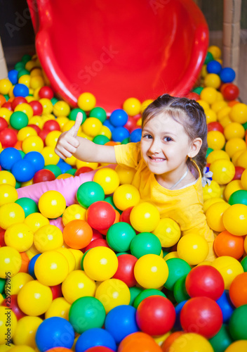 Happy little girl playing and having fun at kindergarten with colorful balls and show thumb up in play center