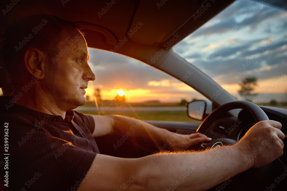 Fototapeta premium man sitting behind the wheel of car during his anticipated travel on summer vacations, man riding in automobile at the countryside with beautiful evening sky and sun rays. Lifestyle and travel concept