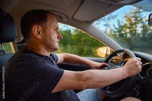 man sitting behind the wheel of car during his anticipated travel on summer vacations, man riding in automobile at the countryside with beautiful evening sky and sun rays. Lifestyle and travel concept