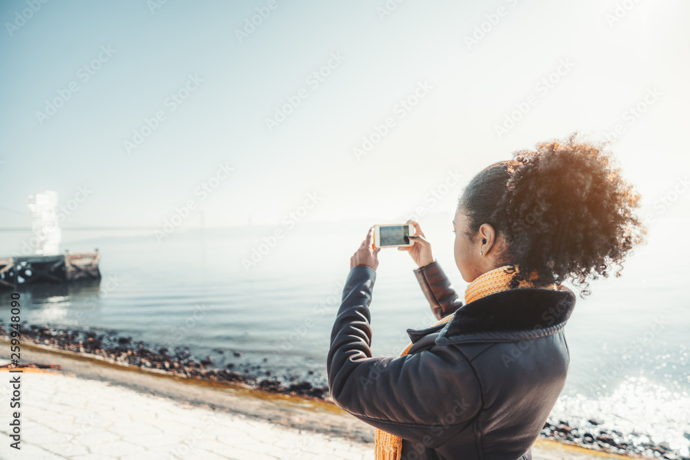 Side view of a curly-hair young African-American female in a warm coat photographing seascape using the camera of her smartphone while standing on the quayside of the river on a sunny spring day