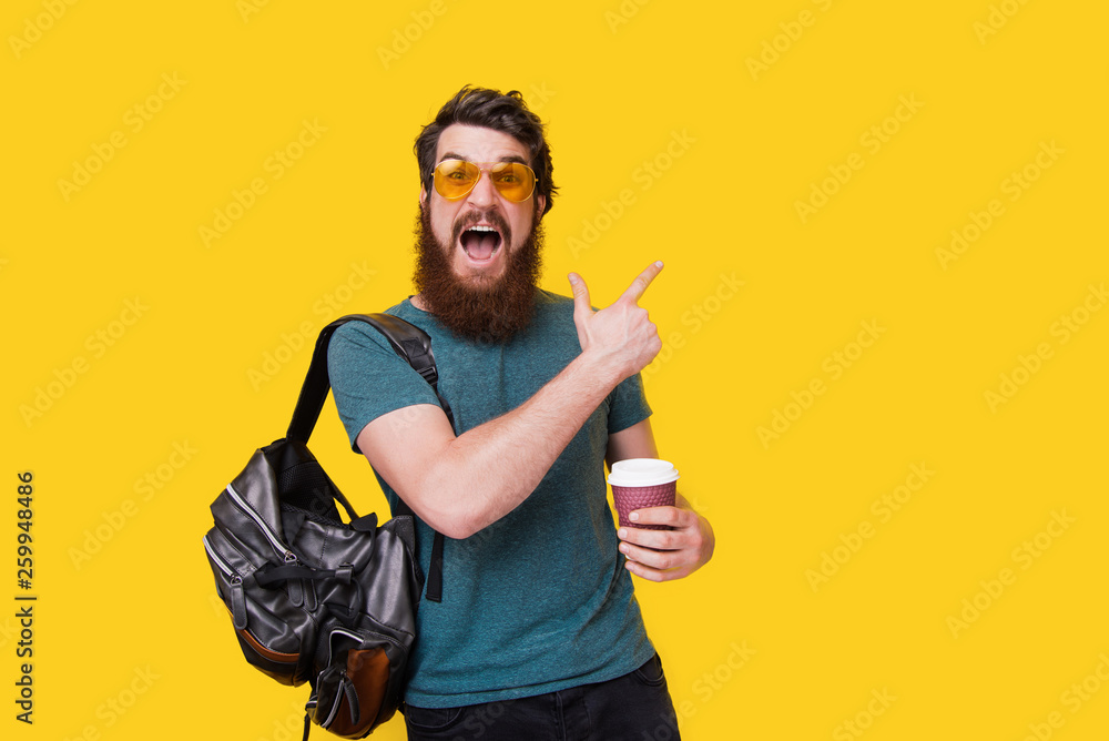 Excited bearded man with stylish eyeglasses, pointing away while holding a cup with coffee on yellow wall background Excited bearded man with stylish eyeglasses, pointing away while holding a cup with coffee on yellow wall background
