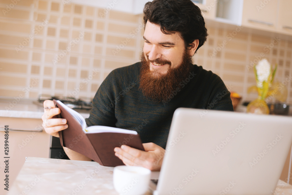 Happy bearded man looking in his notebook sitting in kitchen and work at his business Happy bearded man looking in his notebook sitting in kitchen and work at his business