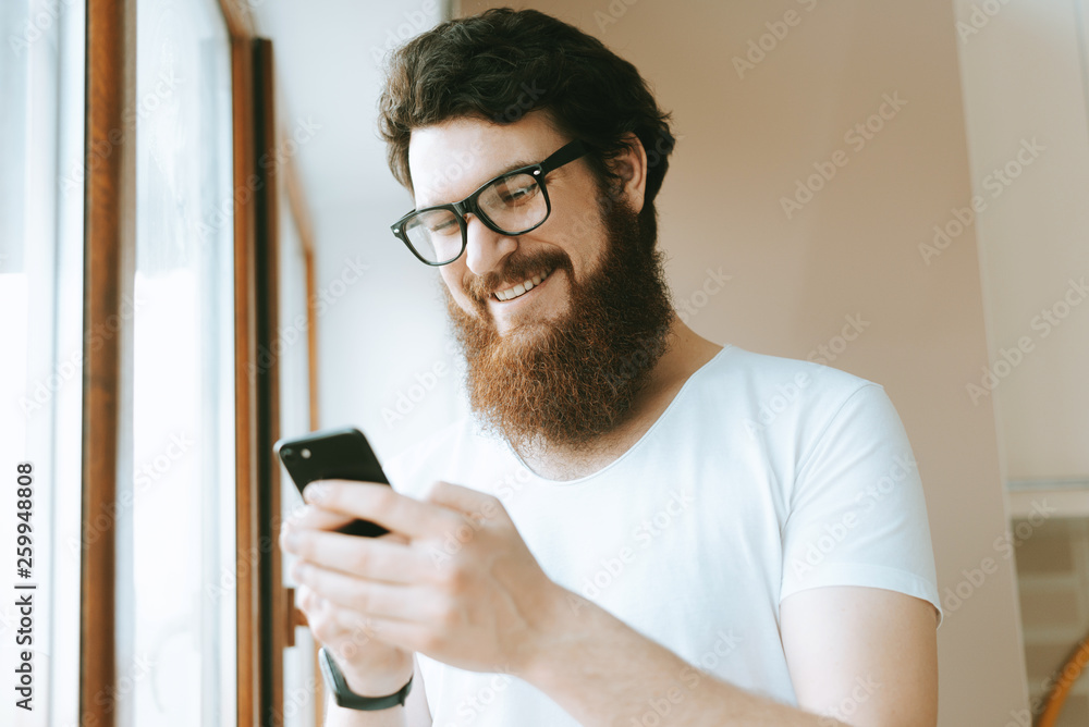 Portrait of smiling bearded man standing near window and using his smaprtphine Portrait of smiling bearded man standing near window and using his smaprtphine
