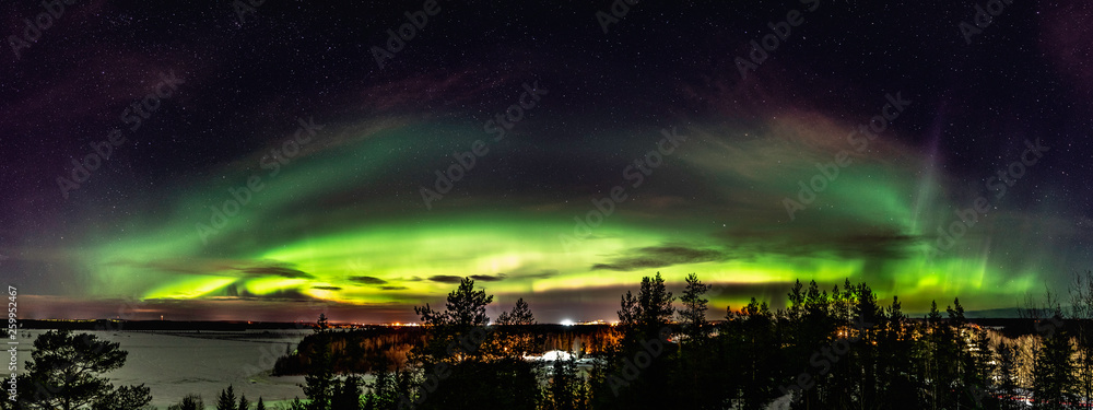 Panoramic view of of brilliant green Aurora. It is curved above horizon line, shining over Scandinavian landscape with frozen river, wild pine tree forest and small villages. Very cold winter night