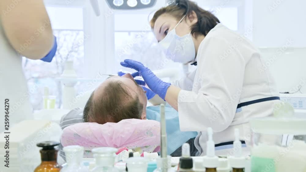 Female dentist with an assistant examines the patient's mouth of a man of an aged man. High key professional work of a dentist in the dental clinic Female dentist with an assistant examines the patient's mouth of a man of an aged man. High key professional work of a dentist in the dental clinic