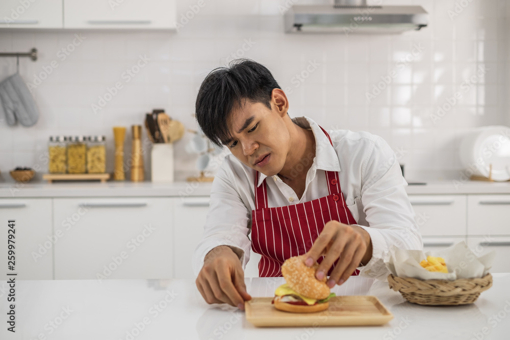 A young Asian man wearing a white shirt and red apron looking at the inside of a burger with curiosity in a white kitchen.