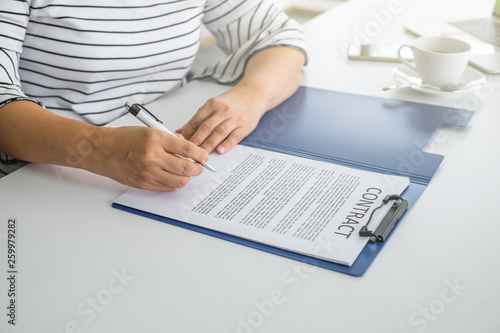 A women hands signing contract in blue folder on top of white table with bright light 