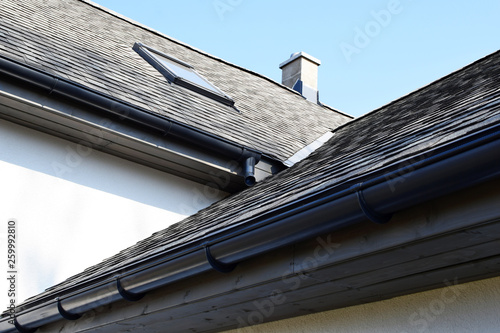Grey asphalt shingles roof construction with ridge ventilation on rooftop, wooden eaves with plastic rain gutter, window and chimney, copy space on white wall or blue sky.