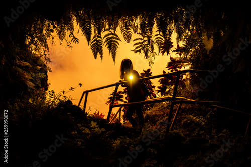 Woman stands near the entrance to the Lava Tube surrounded by lush tropical vegetation and flashes with the light of torch right into the camera. Maui, Hawaii