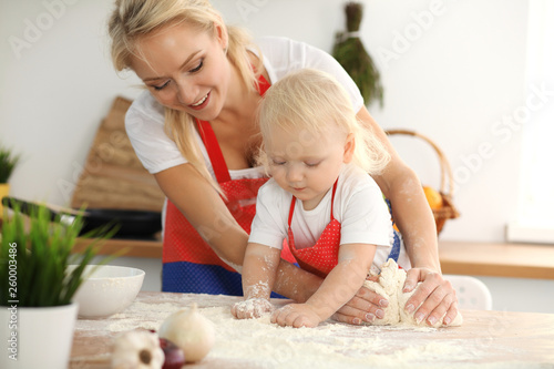 Little girl and her blonde mom in red aprons  playing and laughing while kneading the dough in kitchen. Homemade pastry for bread, pizza or bake cookies. Family fun and cooking concept