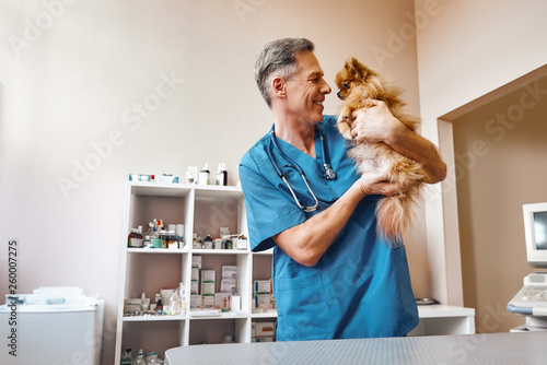 My best part of work! Middle aged positive vet in work uniform talking with small ginger dog while standing at veterinary clinic photo