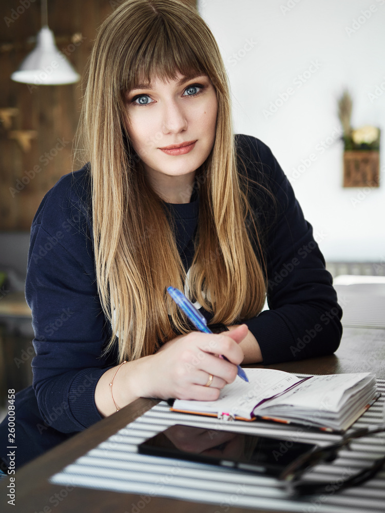 Young serious attractive young woman with pen in hand thinking on plans and writing list to do in notepad enjoying recreation time in stylish coworking space. Thoughtful self employed noting Young serious attractive young woman with pen in hand thinking on plans and writing list to do in notepad enjoying recreation time in stylish coworking space. Thoughtful self employed noting