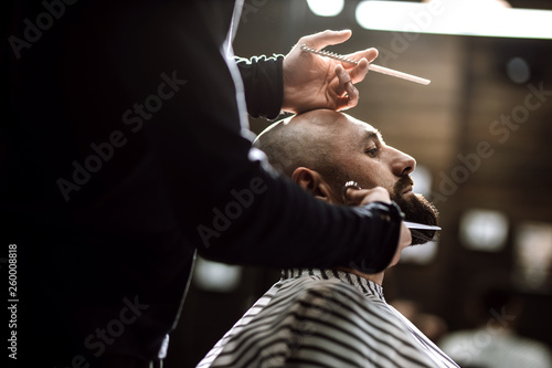 Fashion barber dressed in a black clothes tidies up men's beard  and scissors it in the barbershop photo