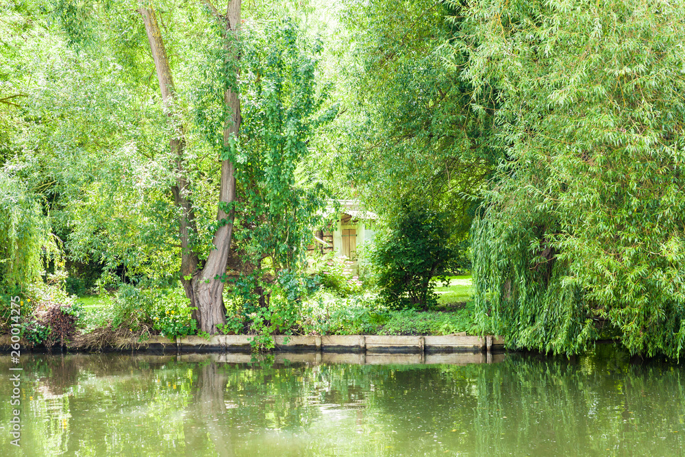 House hidden in vegetation trees and flowers at the edge of a canal in summer. House hidden in vegetation trees and flowers at the edge of a canal in summer.