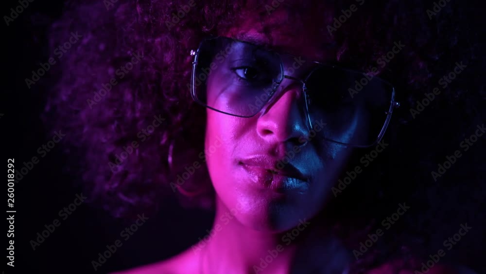 Closeup portrait of trendy black woman with afro hair in neon purple light smiling and looking at camera in studio against dark background. slow motion. Closeup portrait of trendy black woman with afro hair in neon purple light smiling and looking at camera in studio against dark background. slow motion.