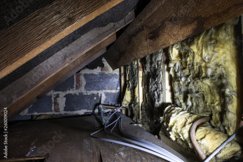 Crawl space under the eves of a house showing old fibreglass insulation, pipework, rafters, breezeblock construction and old boarding.