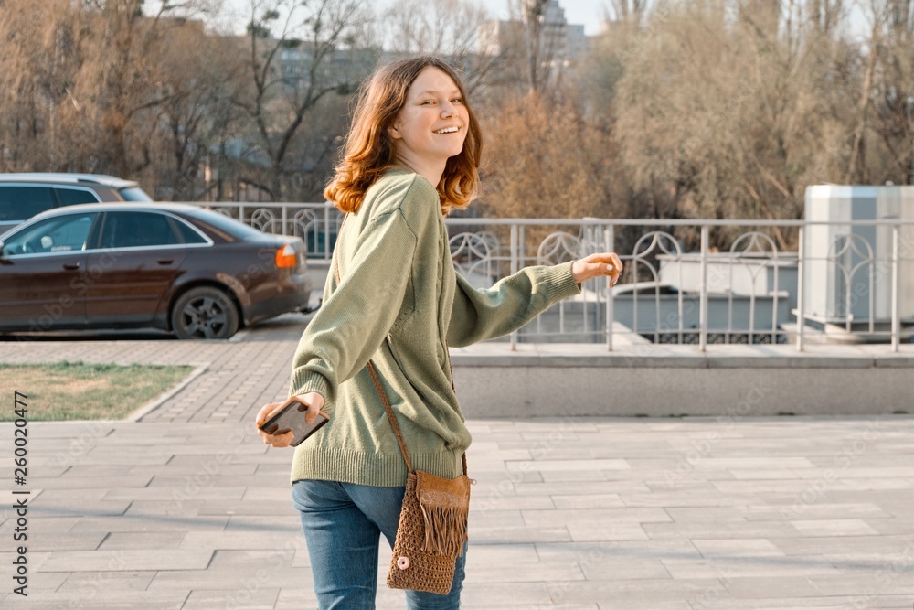 Walking young smiling girl teenager looking in camera through back with brown red hair in green sweater, sunny spring day background, copy space
