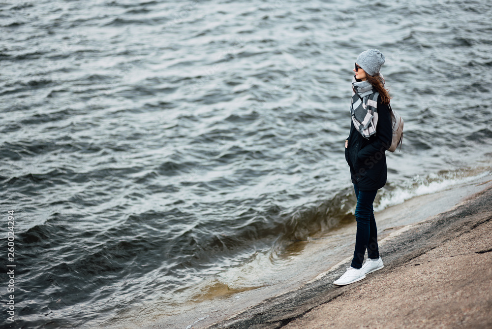 young woman traveler walks along the beach in the fall in windy weather. full length photo of girl in coat, hat, backpack and large scarf goes along the coastline.