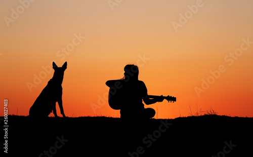 Silhouette girl guitarist on sunset background, silhouette of a dog breed Belgian Shepherd Malinois, happy friends, outdoor