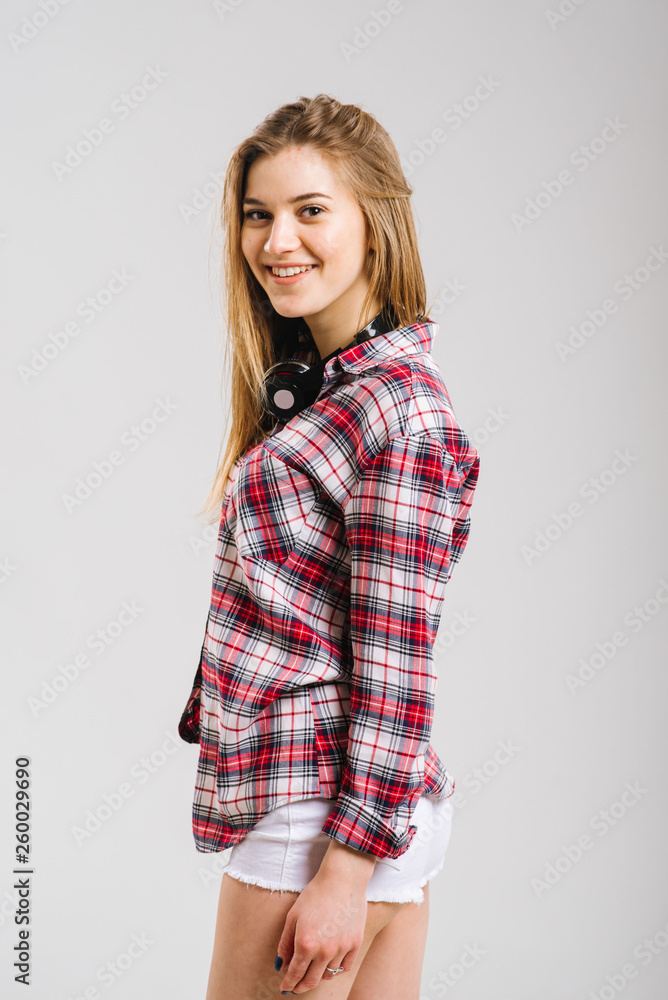 Happy young caucasian woman wearing shirt and shorts with headphones smiling and posing on the grey background Happy young caucasian woman wearing shirt and shorts with headphones smiling and posing on the grey background