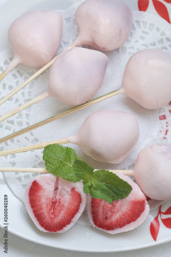 Strawberries in yogurt. Berries are dressed on wooden skewers and dipped in yogurt. Nearby are two halves of dessert. Visible red juicy strawberry flesh. Decorated with mint leaves.