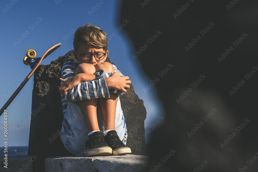 Child alone sitting near the ocean with skateboard. Sad boy crying in a  sunny summer day.