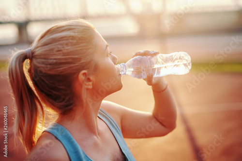 Portrait of young blonde Caucasian woman in sportswear standing on court and drinking water after exercising. Winners never quit and quitters never win. photo