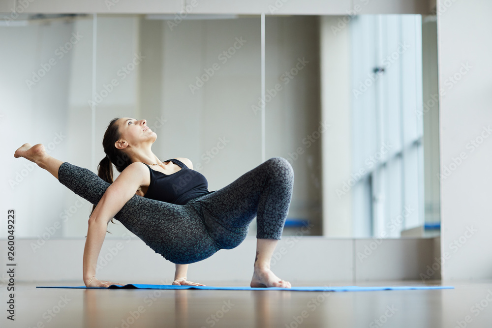 Confident attractive professional yogis in leggings and wifebeater standing  on one foot and raising leg aside through arm while doing modified yoga  crab pose in studio. Photos | Adobe Stock