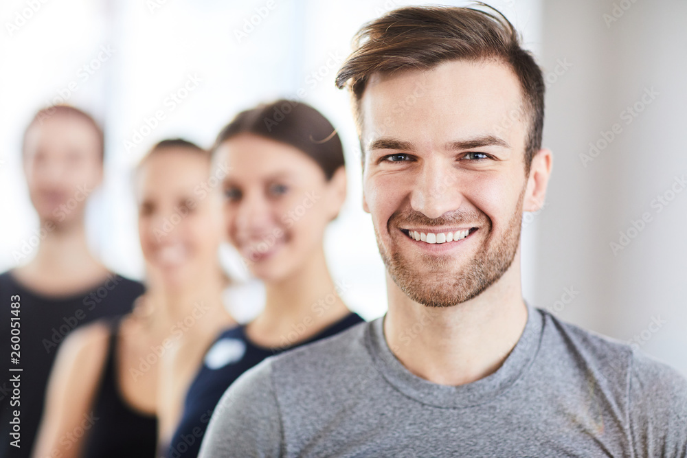 Portrait of cheerful handsome young guy with stubble and fashionable hairstyle smiling at camera while standing in line of fitness trainers Portrait of cheerful handsome young guy with stubble and fashionable hairstyle smiling at camera while standing in line of fitness trainers