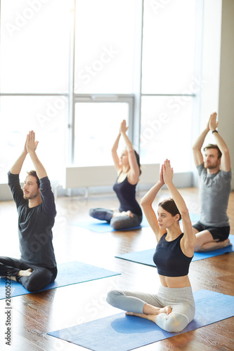 Fototapeta Naklejka Na Ścianę i Meble -  Group of concentrated young people sitting in lotus position on yoga mats in modern room and reaching arms up while doing sun meditation at yoga class.